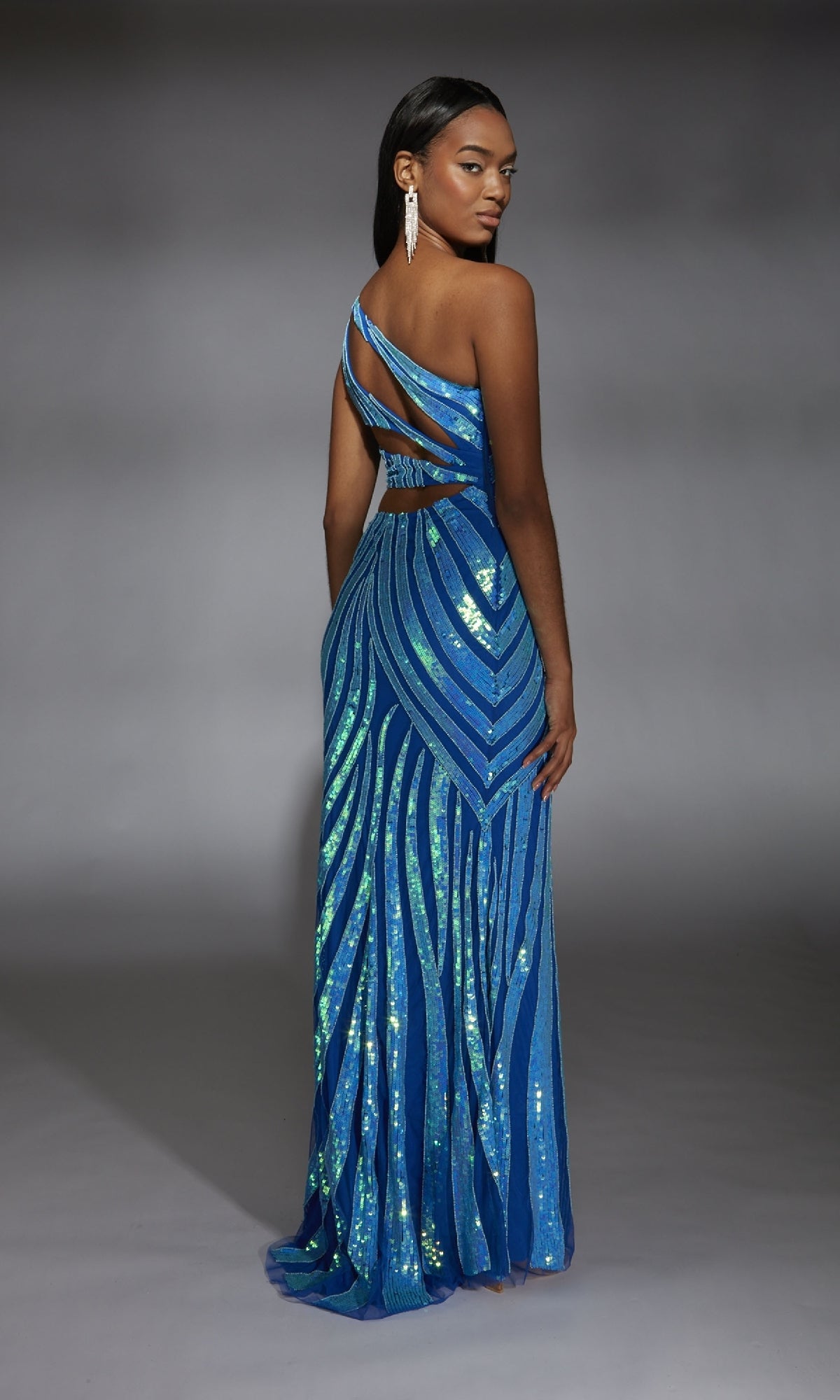 A woman faces away from the camera, wearing the Alyce 61877 long prom dress—a blue sequined, one-shoulder gown with cut-out back details—set against a neutral background.